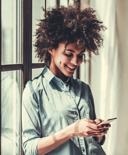 Giovane donna sorridente,  con alle spalle una finestra dalla quale filtra molta luce, consulta la sua posizione da smartphone 