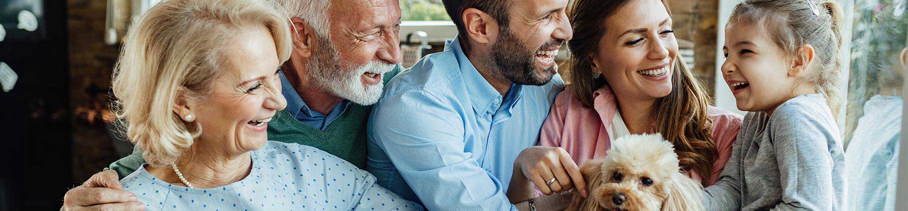 Fotografia di un gruppo familiare sorridente composto da nonni, genitori, una bambina di 5 anni che accarezza un barboncino albicocca. 