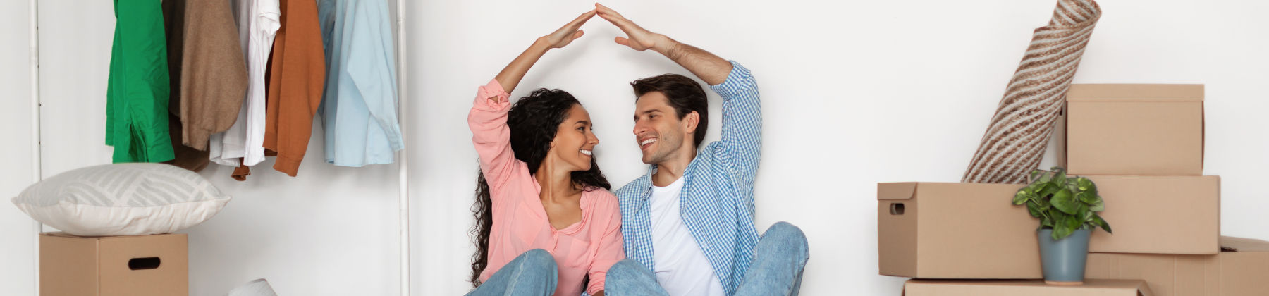 Ragazza e ragazzo tra gli scatoloni del trasloco si guardano sorridenti e uniscono le mani sopra la testa a mo' di tetto.