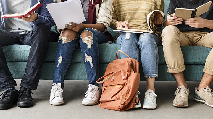 Gruppo di quattro giovani indivdui, di cui non è visibile il volto, sono seduti su divano. Tutti hanno in mano un libro o un quaderno e in atteggiamento amichevole si confrontano sui loro studi.